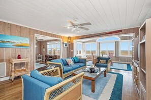 A spacious living room with a large sofa, wicker chairs, a coffee table, and a ceiling fan. - Late for the Sky: Ocean and Waterway Views, Near Beach Access (Caswell Beach)