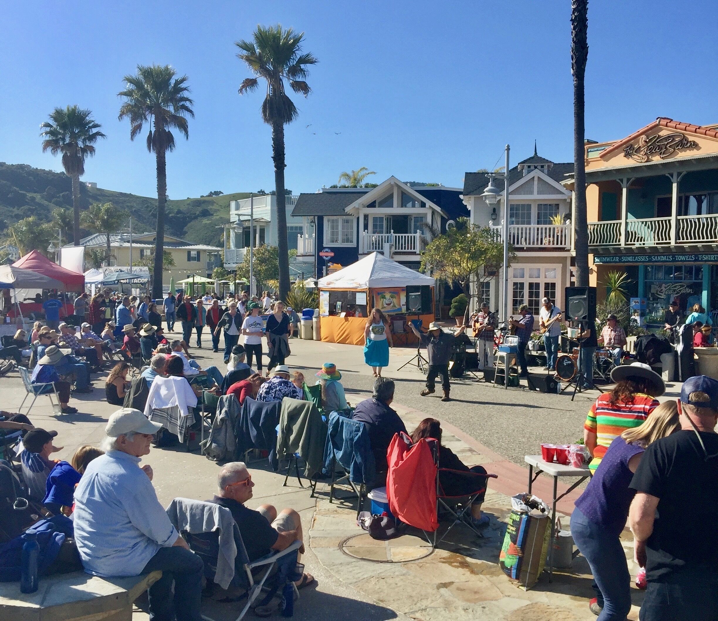A bustling outdoor market with people seated on chairs and others standing, vendors with tents, and a variety of stalls.