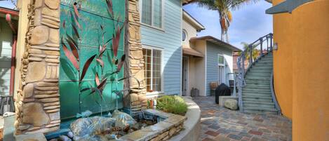 A stone fountain with a water feature, a curved stone wall, and a staircase leading to a door.