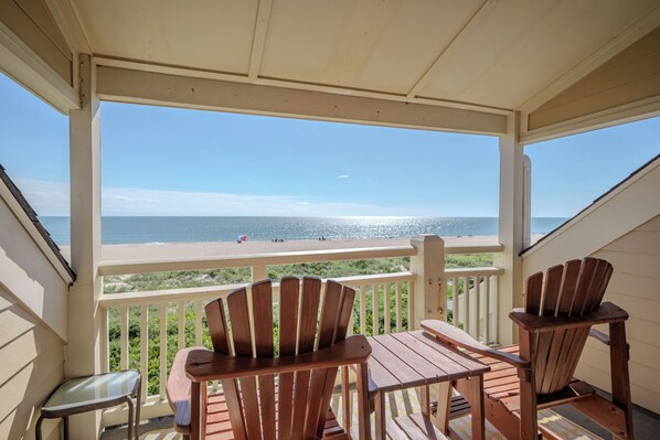 A wooden deck with two Adirondack chairs and a glass table, overlooking a beach with people and a clear sky.
