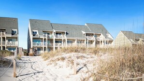 Un hotel fronte spiaggia con numerosi balconi, un vialetto sabbioso che conduce all'ingresso e un cielo azzurro limpido.