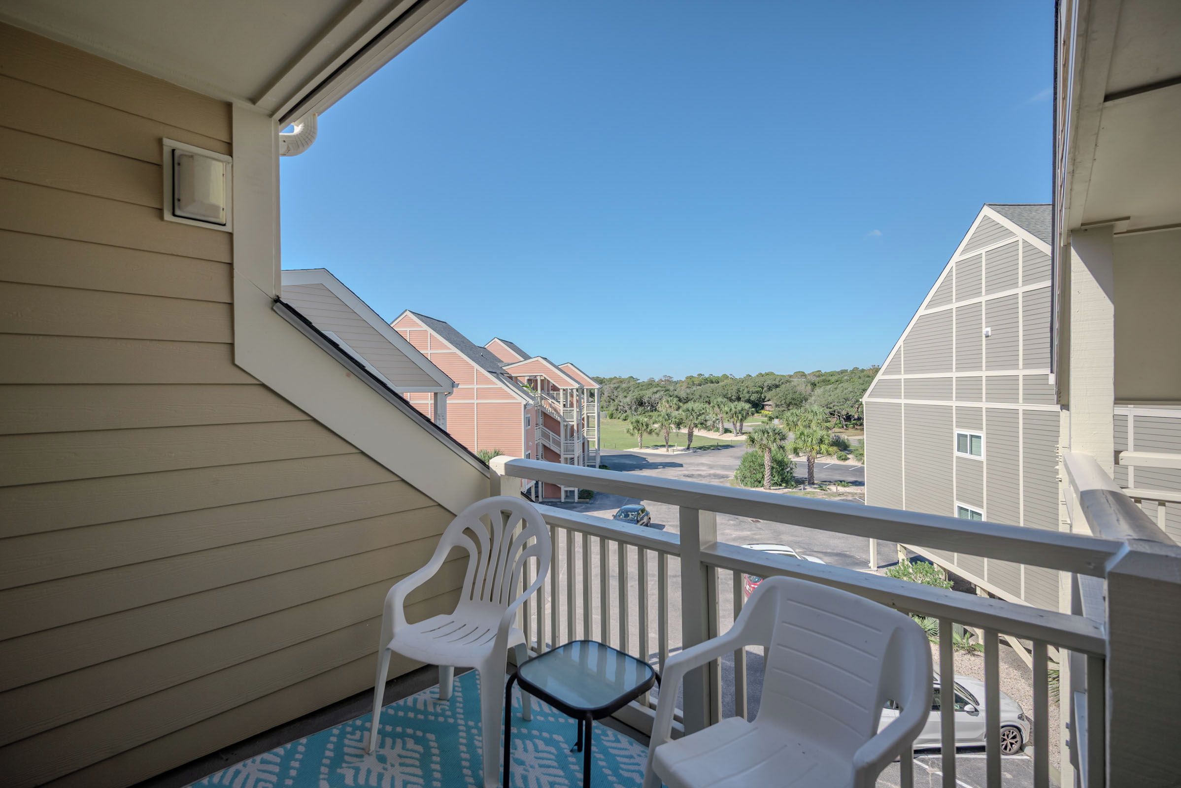 A balcony with two white plastic chairs and a small table, overlooking a parking area and neighboring buildings.