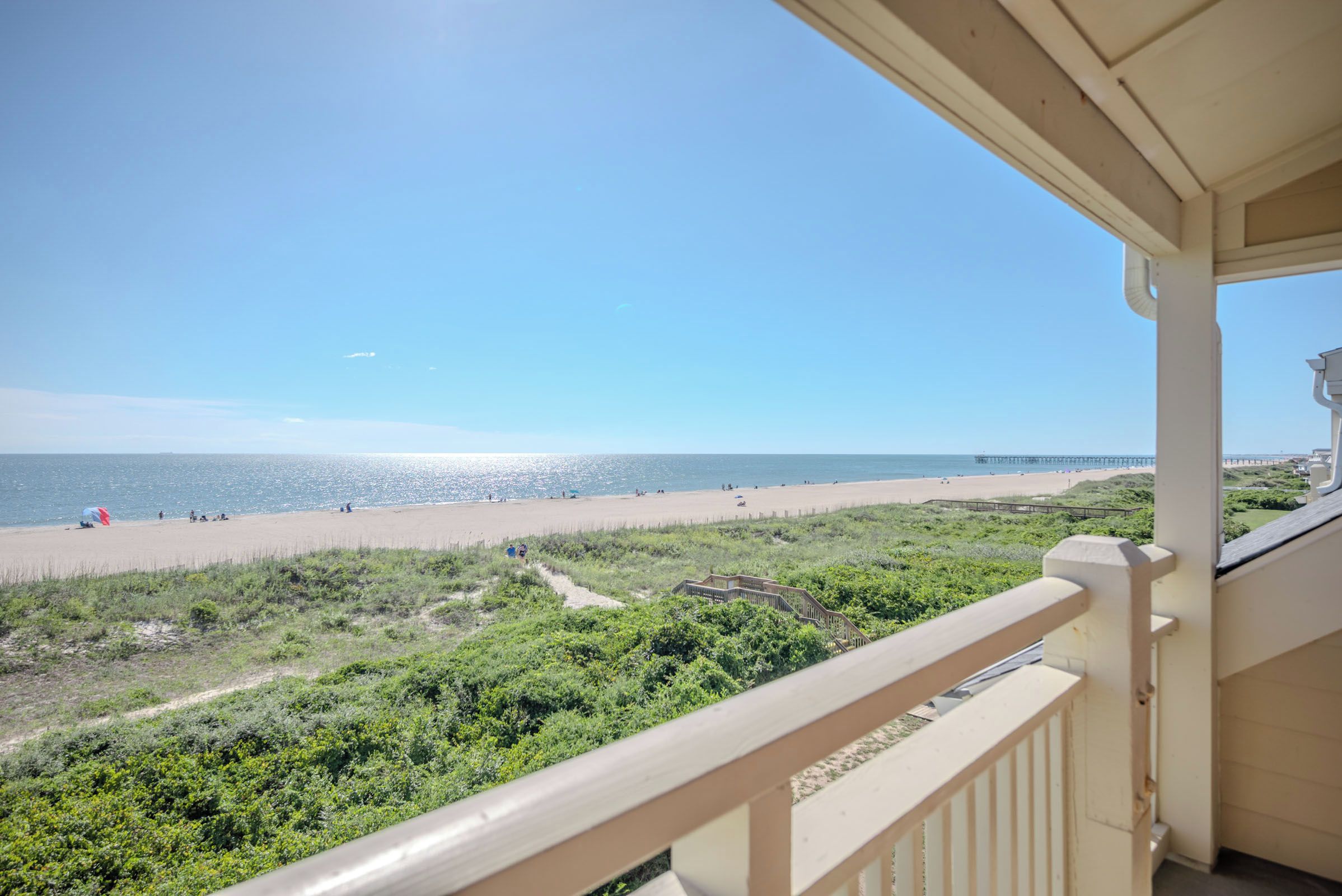 Une vue sur la plage depuis un balcon doté d’une balustrade, avec un ciel dégagé et une jetée visible au loin.