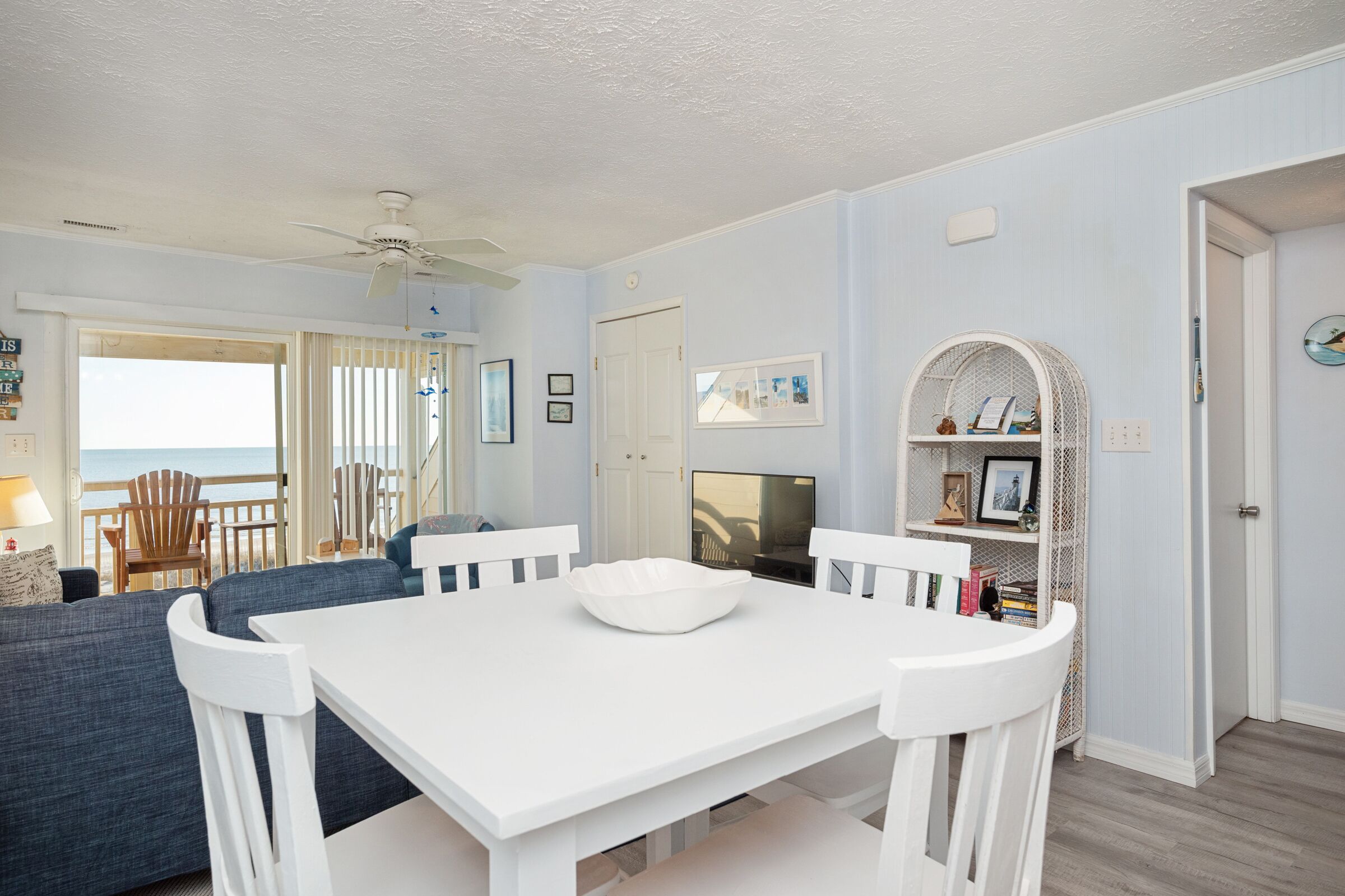 A dining area with a white table and chairs, a blue sofa, and a view of the ocean through a glass door.
