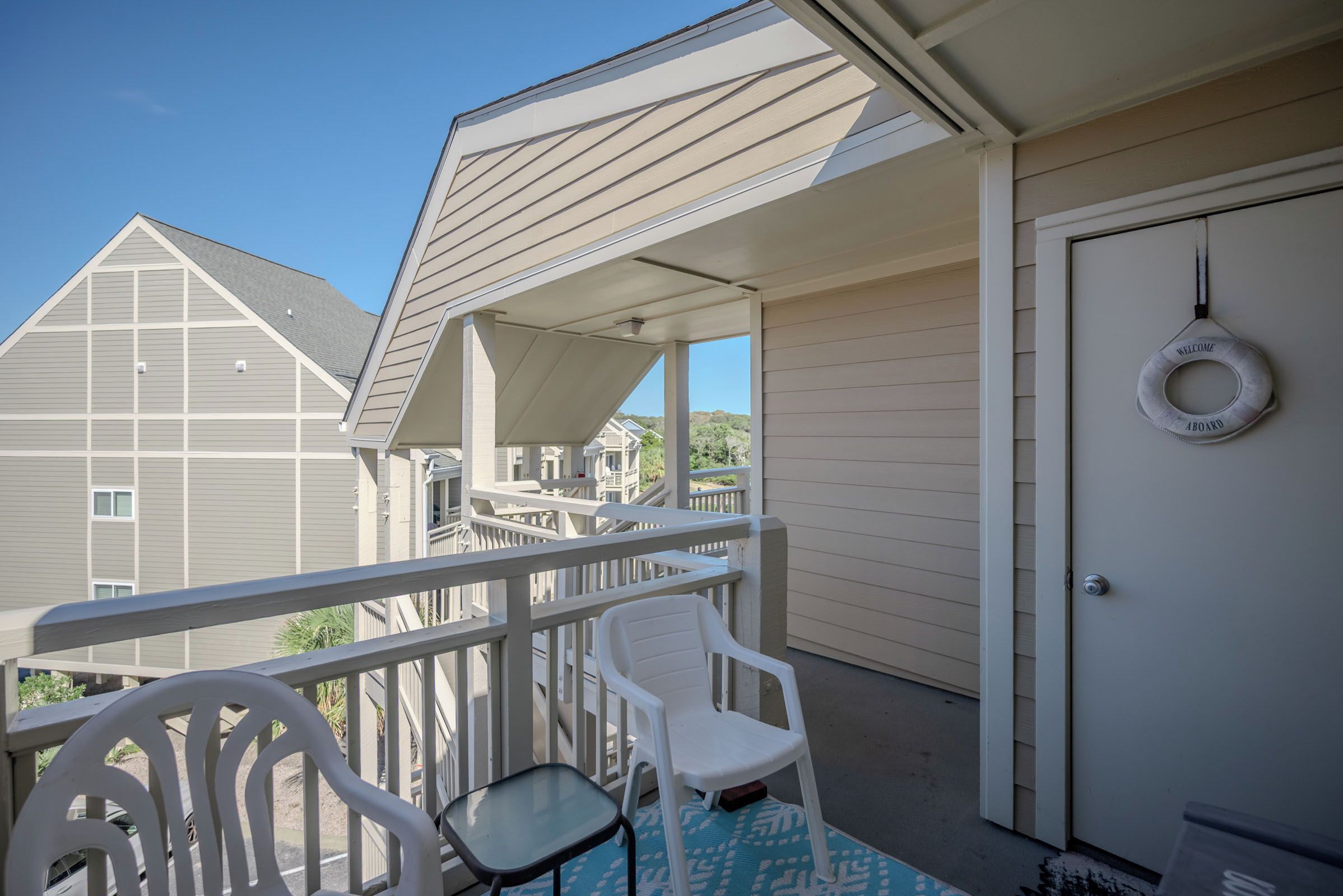 A covered outdoor area with a white chair and a small table, a door with a welcome sign, and a view of neighboring buildings.