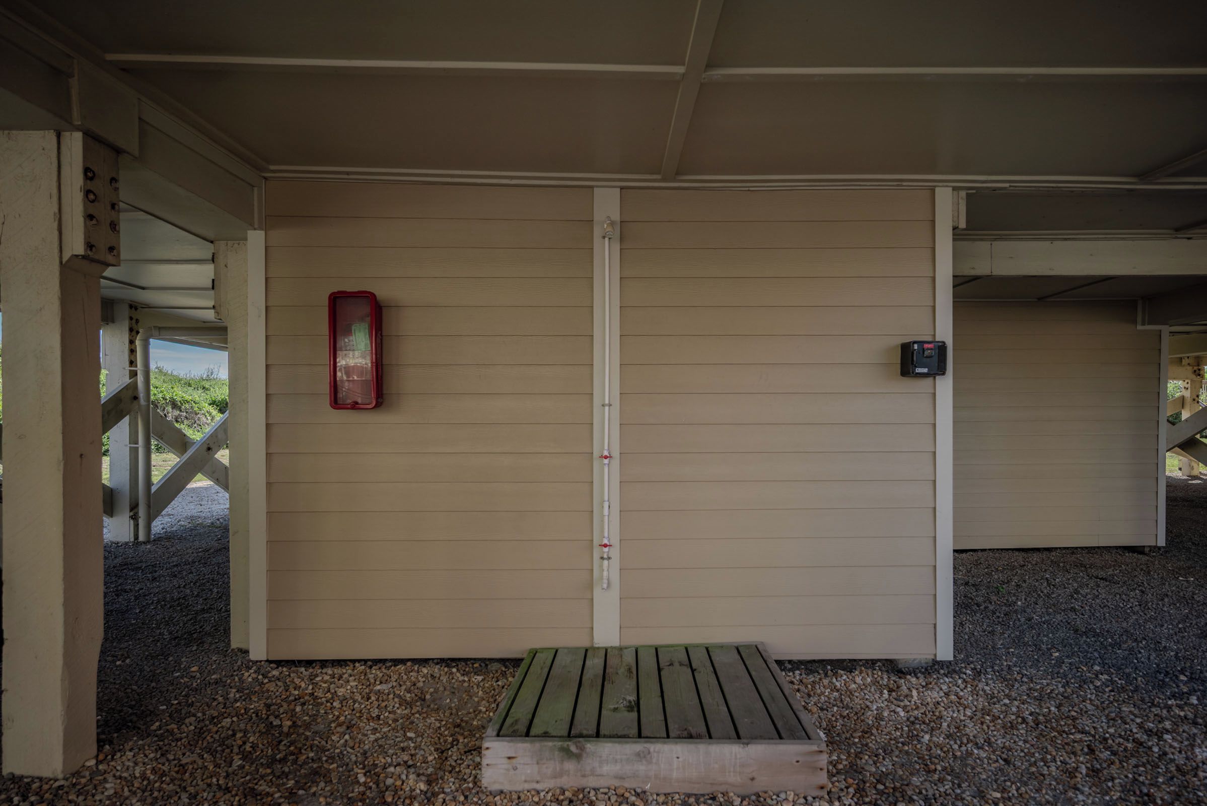 A covered outdoor area with a wooden bench, a red object on the wall, and a black device on the right.