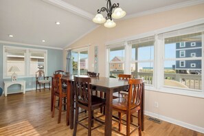 A dining area with a wooden table and chairs, large windows with a view of buildings, and a ceiling light fixture. - This Is It: Great 2nd Row House with Views! (Oak Island)
