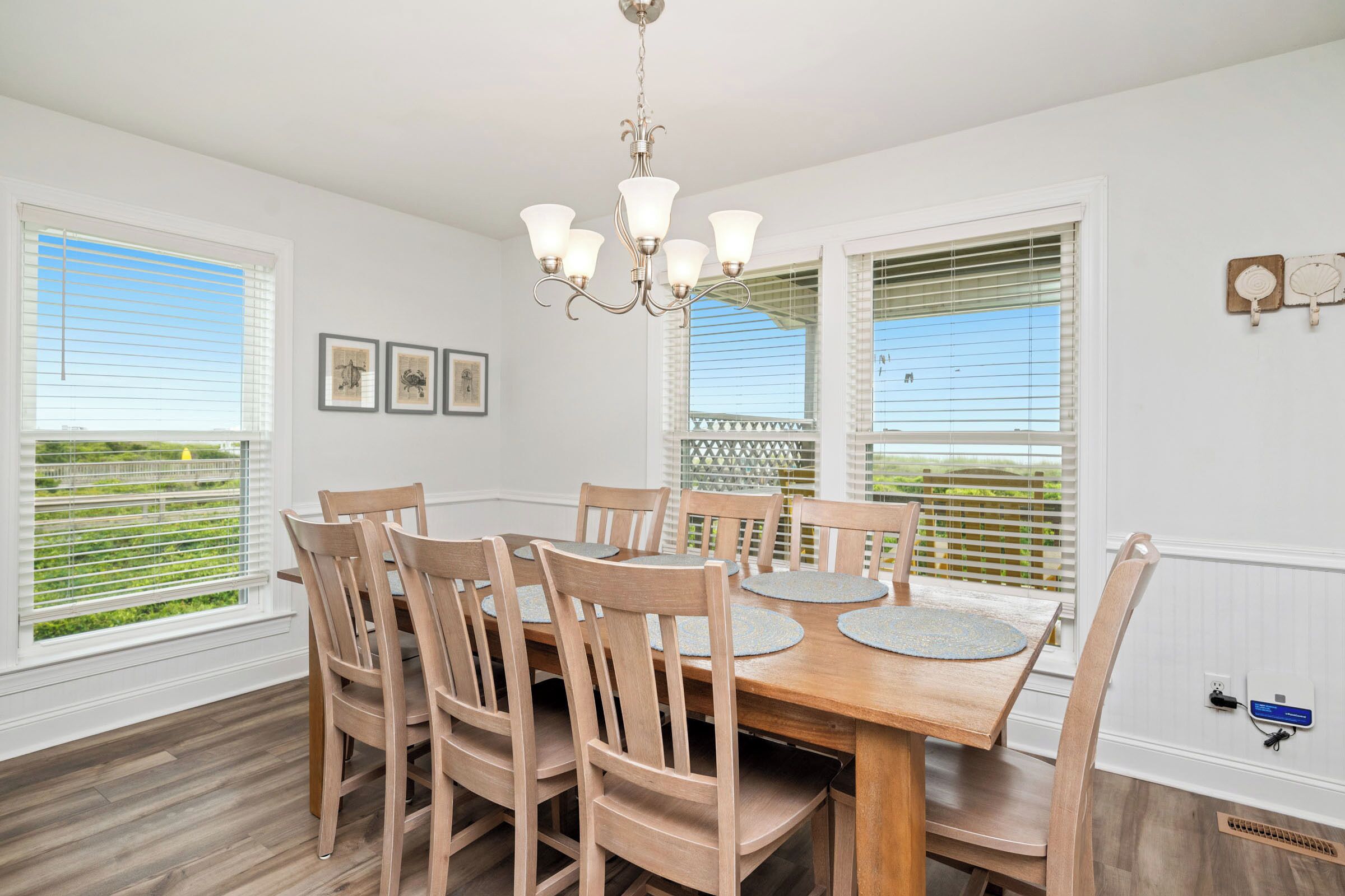 Une salle à manger avec une table et des chaises en bois, un lustre et de grandes fenêtres équipées de stores.