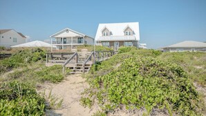 A beachfront property with a wooden deck leading to a house with a white exterior and a balcony. - Linger Longer: Oceanfront with Great Views of Lighthouse (Caswell Beach)
