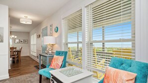 A living room with a sofa, a coffee table, and a lamp. There are windows with blinds, a door, and a dining area in the background. - Linger Longer: Oceanfront with Great Views of Lighthouse (Caswell Beach)
