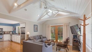 A spacious living area with a sofa, a dining table, and a TV. There is a kitchen with white cabinets and a refrigerator. The room has wooden flooring and a ceiling fan. - Pleasure Point Oasis ~ Family and dog-friendly yard ~ Off-street parking (Santa Cruz)