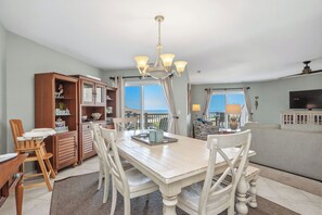 A dining area with a white table, chairs, a chandelier, and a view through large windows. - Our Palm Beach: Spacious Oceanfront Condo with Pool Access (Oak Island)