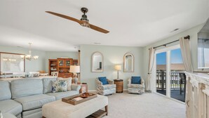 A spacious living room with a ceiling fan, a light blue sofa, a wooden coffee table, and a view of the outside through a large window.