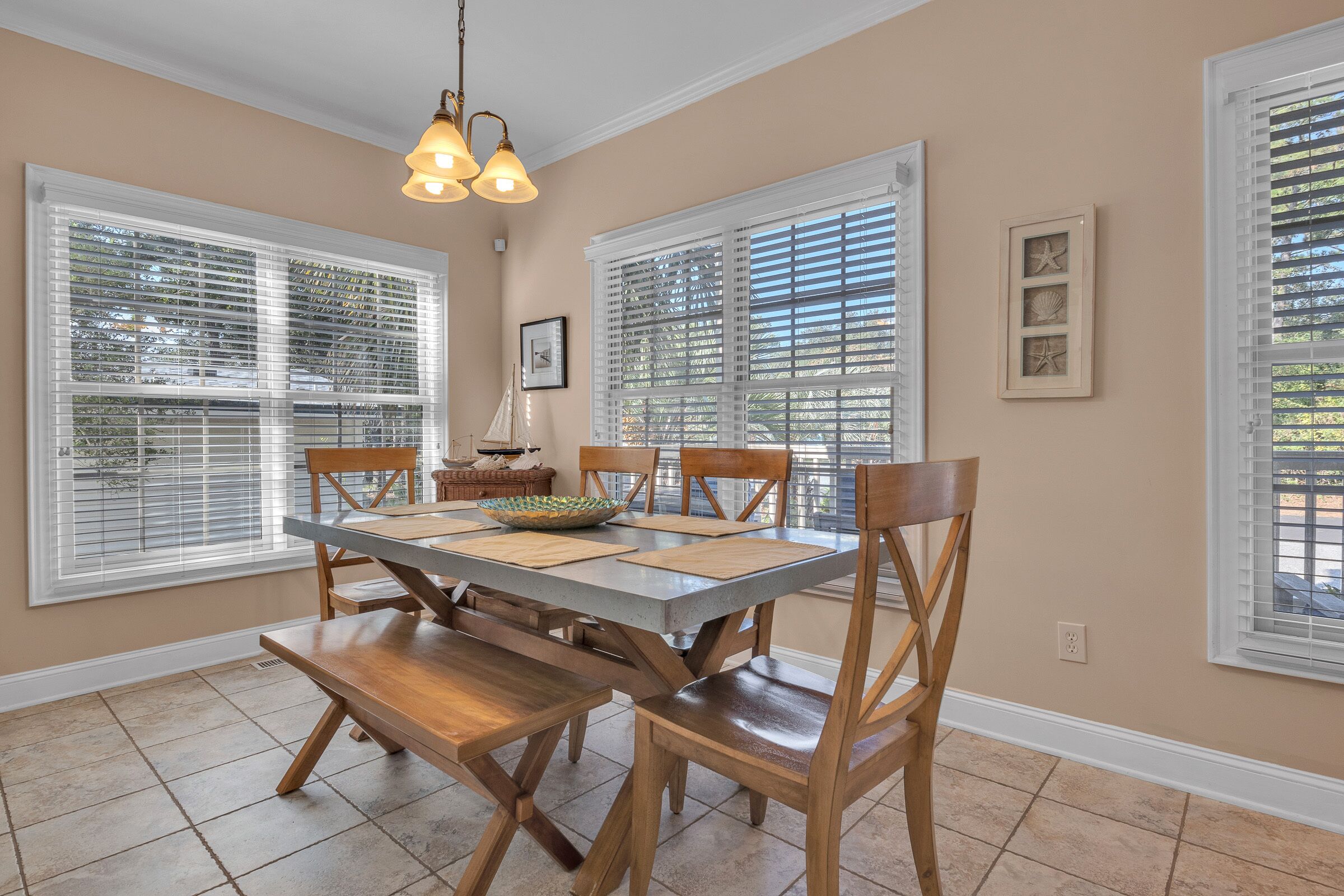 A dining area with a wooden table and chairs, large windows with blinds, and a hanging light fixture.