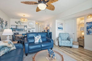 Una sala moderna con un sofá azul, un sillón estampado, un comedor y una cocina al fondo.