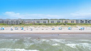 A beachfront hotel with multiple buildings, a sandy beach, and umbrellas. - By The Ocean Blue: Oceanfront Luxury on Caswell Beach with Community Pool (Caswell Beach)