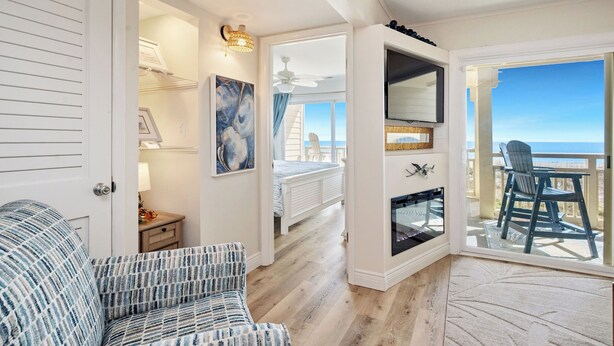A room with a bed, a chair, a fireplace, a TV, and a balcony with a view. - By The Ocean Blue: Oceanfront Luxury on Caswell Beach with Community Pool (Caswell Beach)