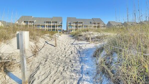 A sandy path leading to a beachfront hotel with multiple balconies and a clear blue sky. - By The Ocean Blue: Oceanfront Luxury on Caswell Beach with Community Pool (Caswell Beach)