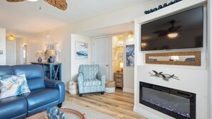 A living room with a blue sofa, a patterned armchair, a fireplace, and a large flat-screen TV mounted above it. - By The Ocean Blue: Oceanfront Luxury on Caswell Beach with Community Pool (Caswell Beach)