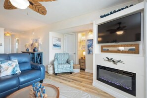 Un salon avec un canapé bleu, un fauteuil à motifs, une cheminée et un grand téléviseur à écran plat fixé au-dessus.