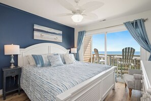 Et soveværelse med seng, sengeborde, loftventilator, udsigt til stranden og en balkon med stole.