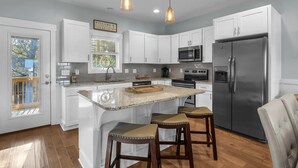 A modern kitchen with white cabinets, a stainless steel refrigerator, and a large island with stools. - All Summer Long: Modern Home located in the Woodlands and Waterway area of Oak I (Oak Island)