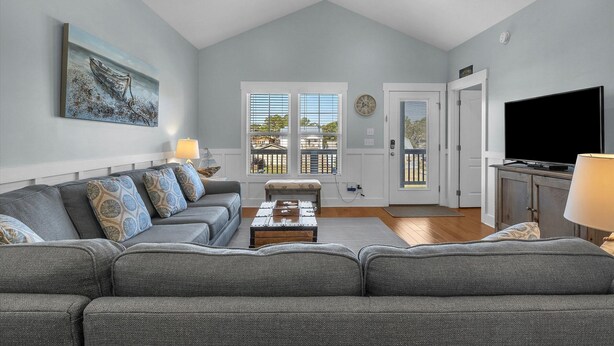 A spacious living room with a gray sectional sofa, a wooden coffee table, and a flat-screen TV. There are two windows with blinds, a ceiling fan, and a door leading to another room. - All Summer Long: Modern Home located in the Woodlands and Waterway area of Oak I (Oak Island)