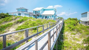 A coastal house with a wooden deck leading to a large house with multiple levels and a balcony. - Beach Buzz: Oceanfront with Expansive Beach and Sound Side Decks (Caswell Beach)
