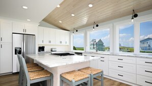 A modern kitchen with white cabinetry, a large island with bar stools, and a view of houses through large windows. - Beach Buzz: Oceanfront with Expansive Beach and Sound Side Decks (Caswell Beach)