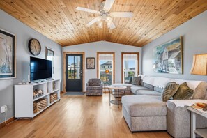A spacious living room with a ceiling fan, a flat-screen TV, a sofa, a chair, and a small table. - Beach Treat: Coastal Chic Home with Spacious Deck, Close to Beach Access (Oak Island)
