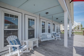 A covered porch with white Adirondack chairs and a small table. - Beach Treat: Coastal Chic Home with Spacious Deck, Close to Beach Access (Oak Island)