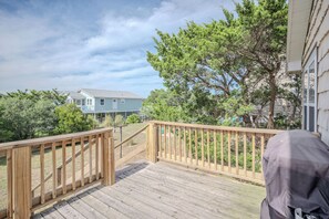 Eine Holzterrasse mit Geländer, einem Grill und Blick auf ein Haus sowie Bäume im Hintergrund.
