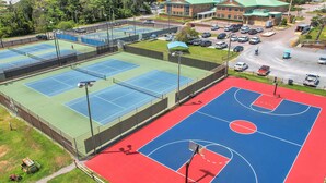 Instalación deportiva al aire libre con varias canchas de tenis y baloncesto, rodeada de un estacionamiento y un edificio al fondo.