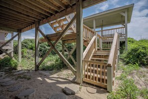 Una estructura de madera con una escalera que lleva a una zona cubierta, rodeada de arena y vegetación.
