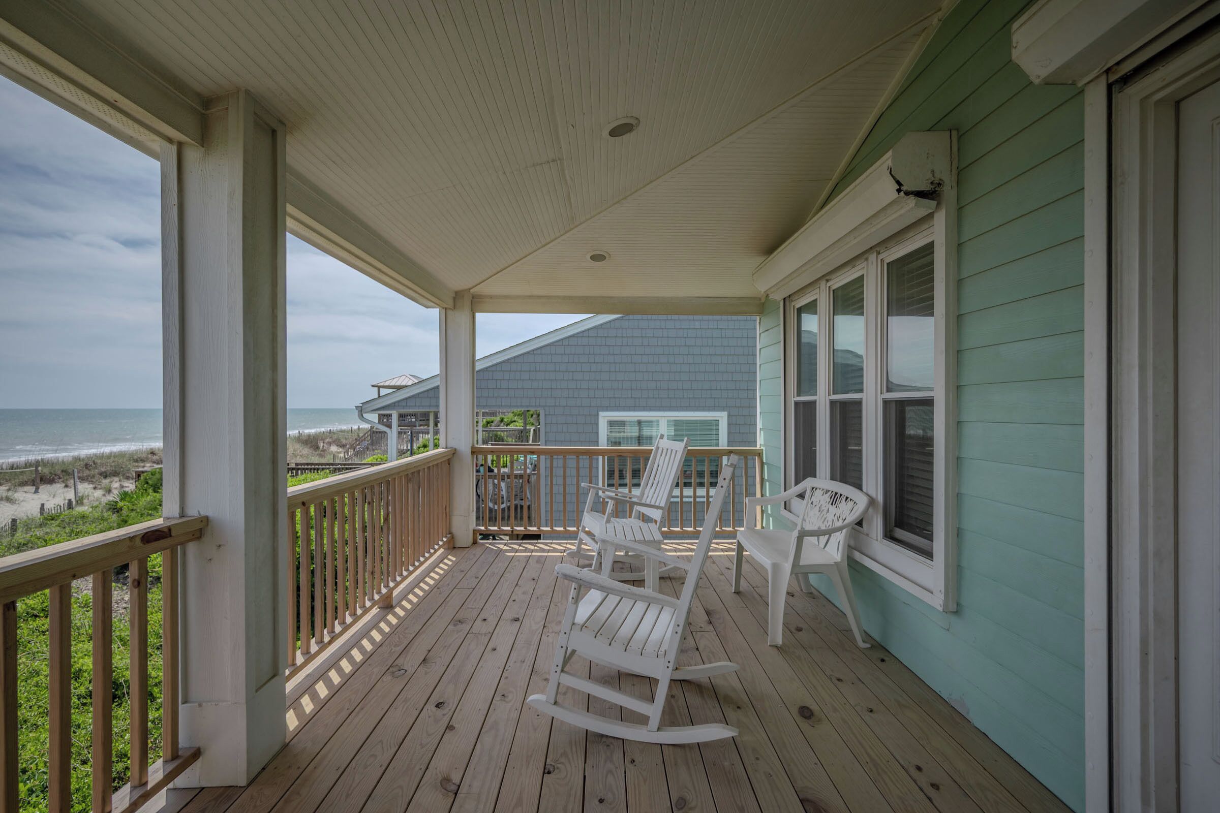 Eine überdachte Veranda mit Schaukelstühlen, Blick auf den Strand und ein Haus mit blauer Fassade.