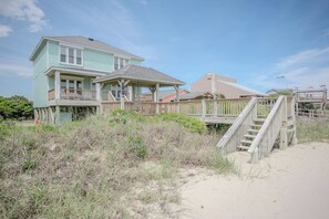 Una casa frente a la playa con un muelle de madera, un porche cubierto y una escalera que baja a la playa.