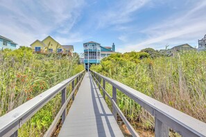 A wooden boardwalk leading to a blue building with a balcony, surrounded by tall grasses and other vegetation.