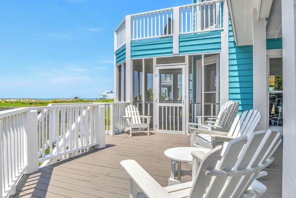 A deck with white chairs and a table, overlooking a beach.