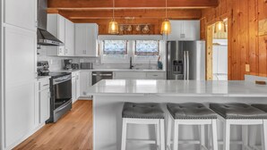 A modern kitchen with white cabinets, stainless steel appliances, and a wooden ceiling. There are three pendant lights hanging above the island. - Pelican Crossing: Oceanfront Home w/Spiral Staircase (Oak Island)
