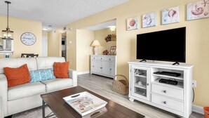 A living room with a white sofa, brown cushions, a coffee table with a magazine, a TV on a white cabinet, and a wall clock. - Balcony At The Beach: Oceanfront Condo with Community Pool (Caswell Beach)
