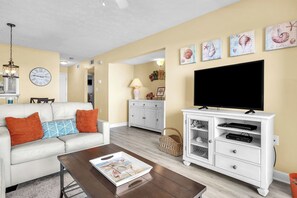Ein Wohnzimmer mit einem weißen Sofa, braunen Kissen, einem Couchtisch mit einer Zeitschrift, einem Fernseher auf einem weißen Schrank und einer Wanduhr.