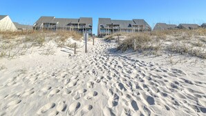 A sandy beach path leading to a row of modern beach houses with multiple balconies. - Balcony At The Beach: Oceanfront Condo with Community Pool (Caswell Beach)
