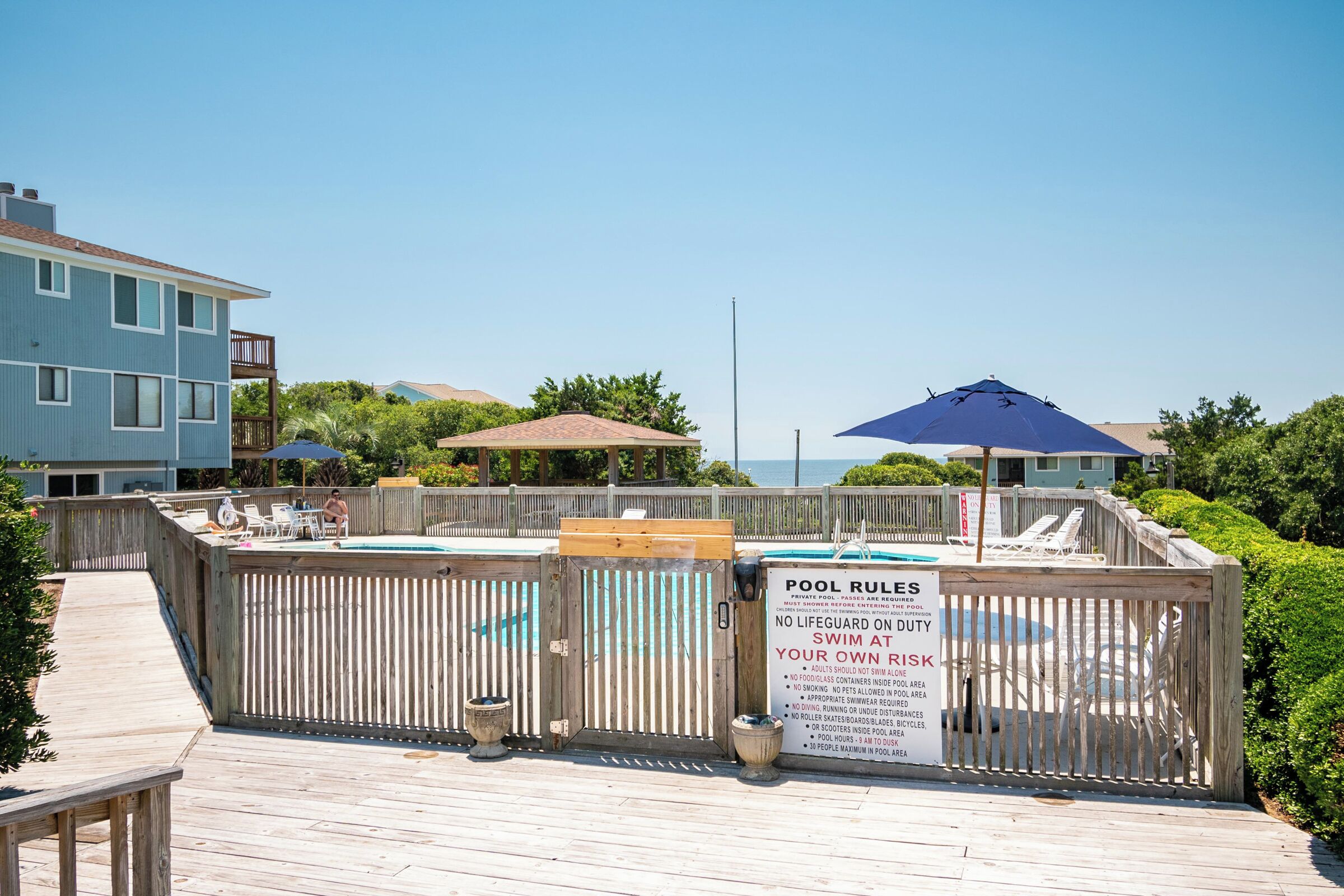 A pool area with a wooden fence, a sign with pool rules, and a view of a building and trees.