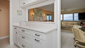 Een moderne keuken met witte kasten, een groot eiland en uitzicht op de woonkamer met een bank en rieten stoelen.