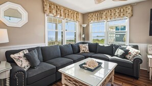 A living room with a sectional sofa, a glass coffee table, and large windows with curtains. - 14 Us: Oceanfront Home with Kayaks, Horseshoe Pit and More Close to Pier (Oak Island)