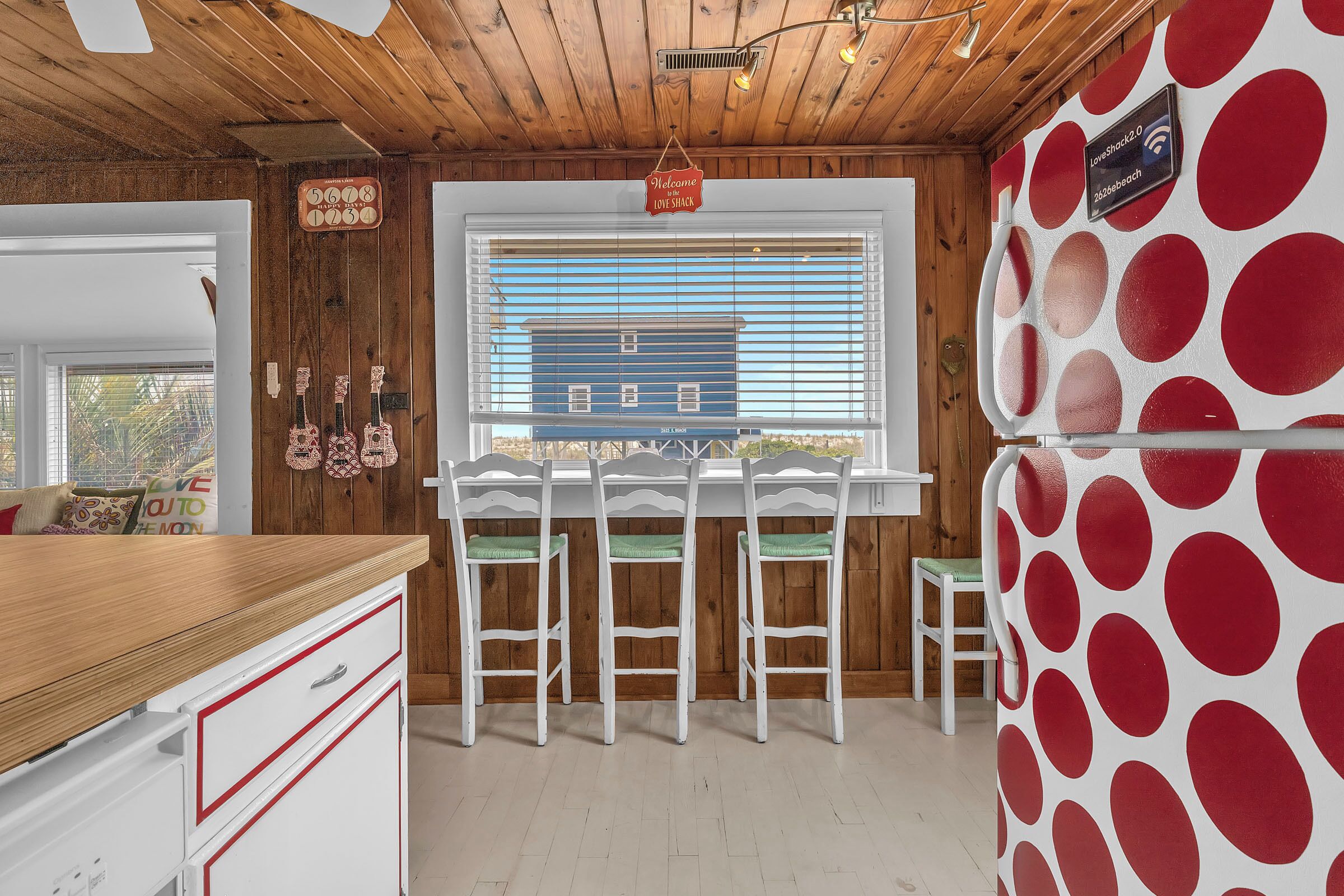 A kitchen with wooden walls, a window with blinds, a refrigerator with a polka dot design, and a bar area with white chairs.