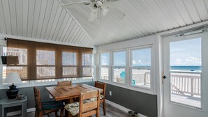 Une chambre située en bord de mer, avec une table et des chaises en bois, de grandes fenêtres donnant sur la plage, un ventilateur de plafond et une porte menant à un balcon.