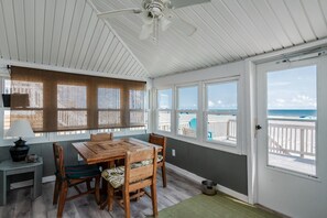 Una stanza sul mare con un tavolo e sedie in legno, ampie finestre vista spiaggia, una ventola a soffitto e una porta che dà su un balcone.