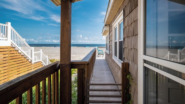 Une terrasse en bois donnant sur la plage, une maison en bois avec de grandes fenêtres et un ciel dégagé.