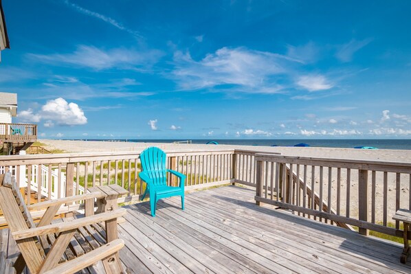 A wooden deck with a turquoise Adirondack chair, overlooking a beach with a wooden railing and a clear blue sky.